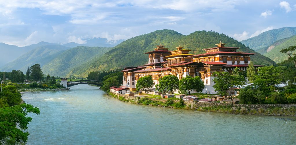 Bhutan's Punakha Dzong, a large white and orange building, sits beside a wide river with mountains in the background.