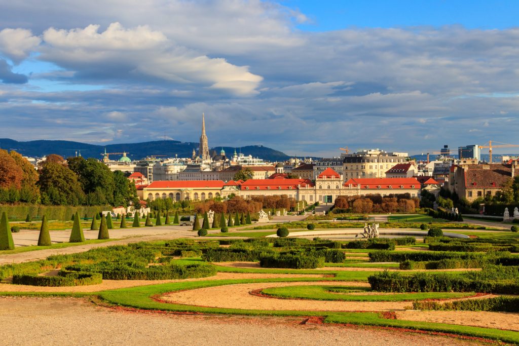 Wide view of Belvedere Palace gardens, Vienna, with city skyline, blue sky, and manicured hedges.