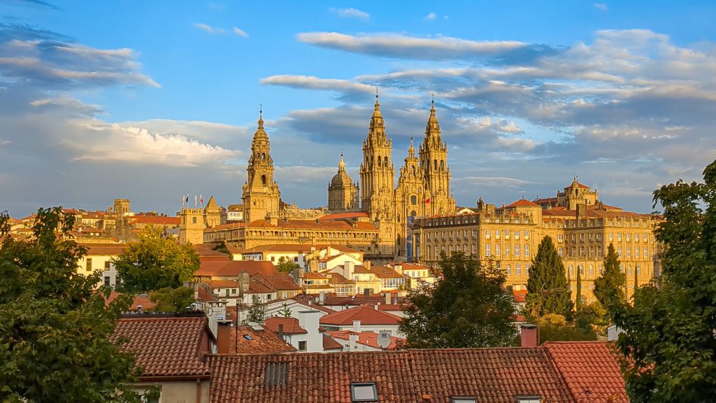 Scenic view of Santiago de Compostela Cathedral with golden light, red rooftops, and blue sky with clouds.