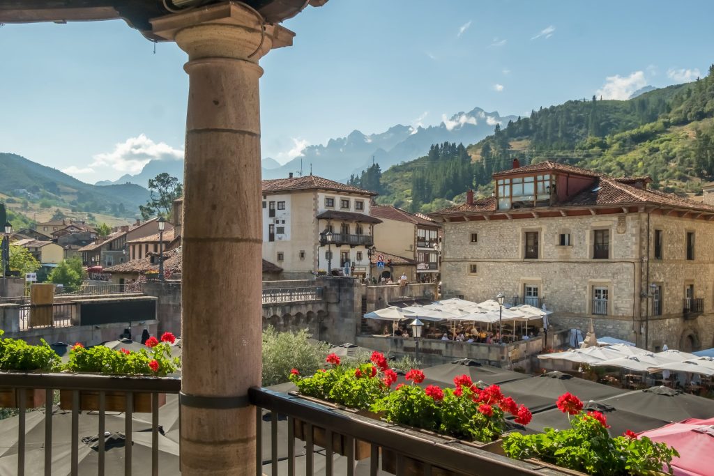 View from balcony with red geraniums overlooking Potes, Spain. Stone buildings, mountains, and people dining under umbrellas.