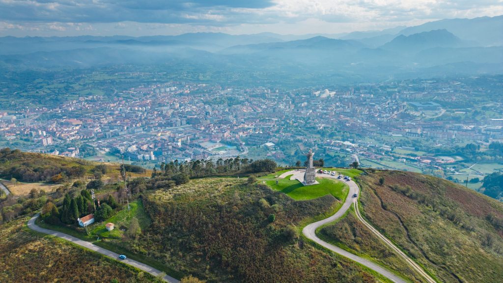 Aerial view of Oviedo, Spain, with a monument on Monte Naranco, winding roads, and distant mountains.