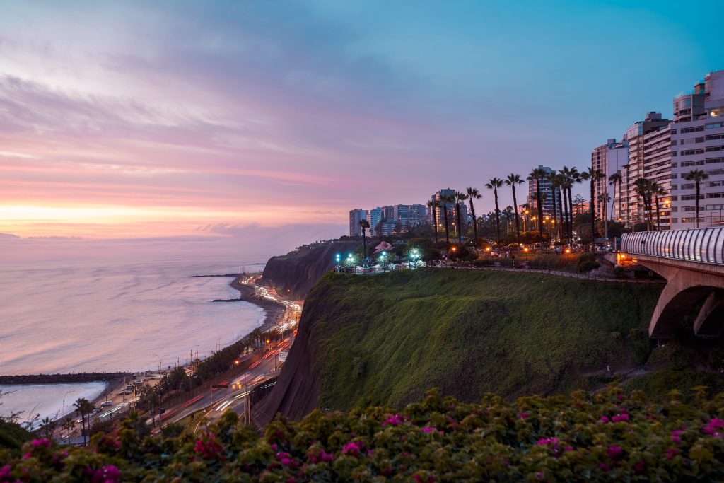 Coastal cityscape at sunset featuring the Miraflores district in Lima, Peru, with ocean, cliffs, buildings, and colorful sky.