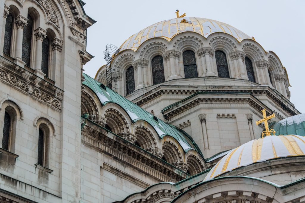 Ornate white stone cathedral with gold domes and crosses, intricate carvings, and arched windows against a gray sky.