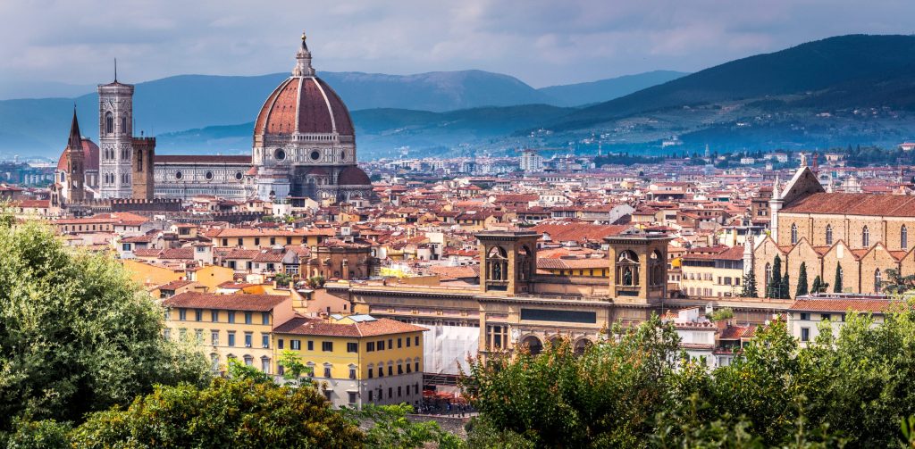 Panoramic view of Florence, Italy featuring the Duomo, cityscape, terracotta rooftops, and distant mountains under a cloudy sky.