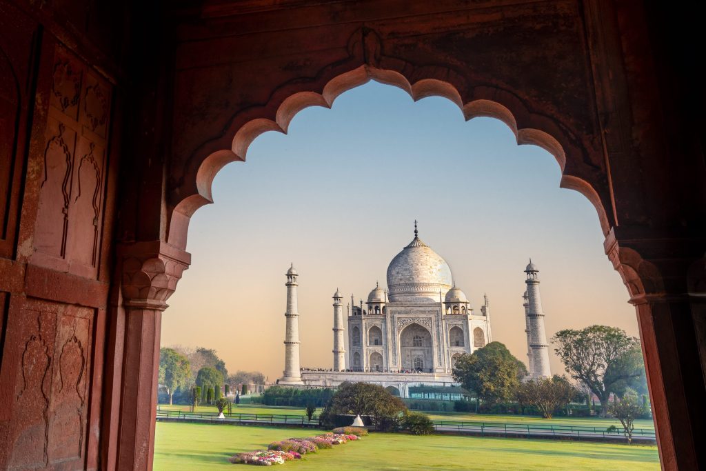 The Taj Mahal is viewed through a red sandstone archway, with green lawns and trees in the foreground.