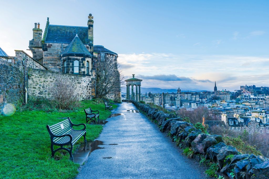 Scenic view of Edinburgh from Calton Hill, featuring cityscape, monument, benches, and wet path.