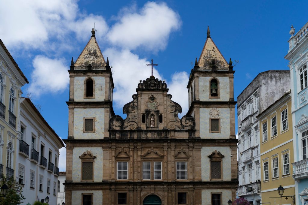 Ornate baroque church facade in Salvador, Brazil, with twin towers, cross, and colorful colonial buildings under a blue sky.
