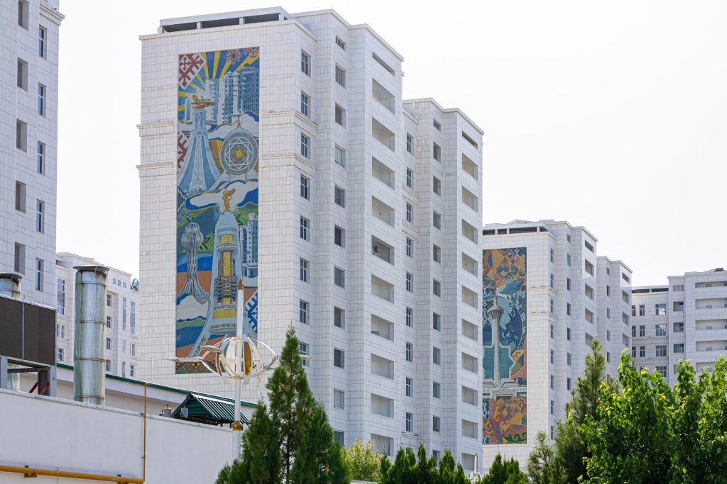 Tall white buildings in Ashgabat, Turkmenistan feature colorful mosaic artwork, viewed through green trees.