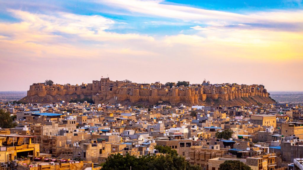 Jaisalmer Fort, a large sandstone citadel, towers over a dense cityscape in Rajasthan, India, under a colorful sunset sky.
