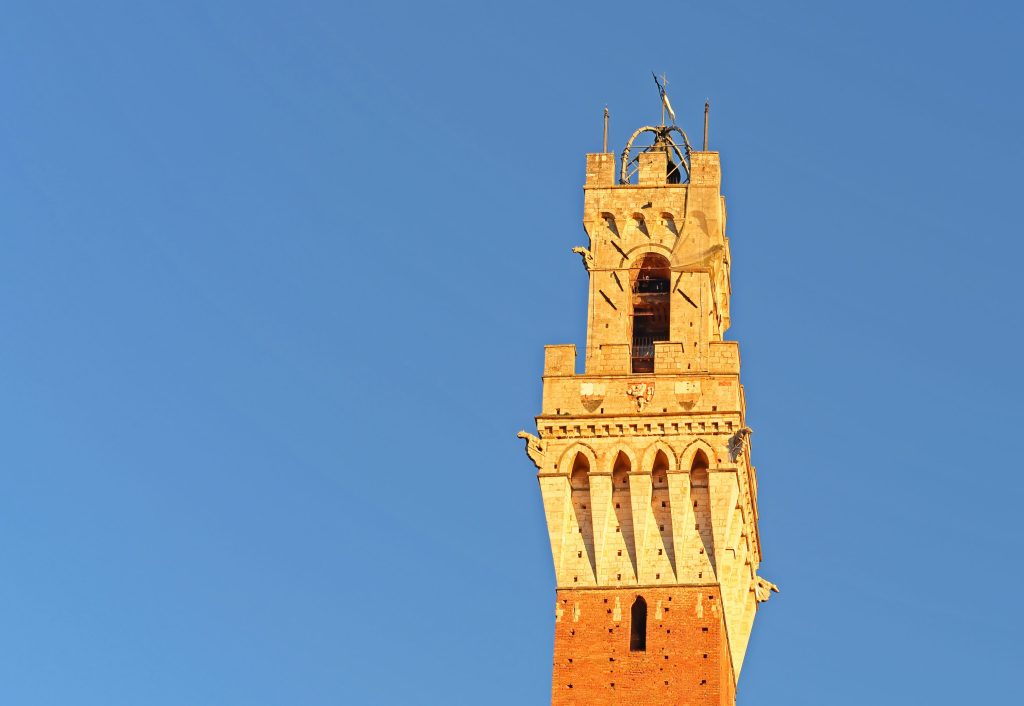 Tall brick and stone tower in Siena, Italy, with blue sky background. The tower has arched windows and decorative elements.