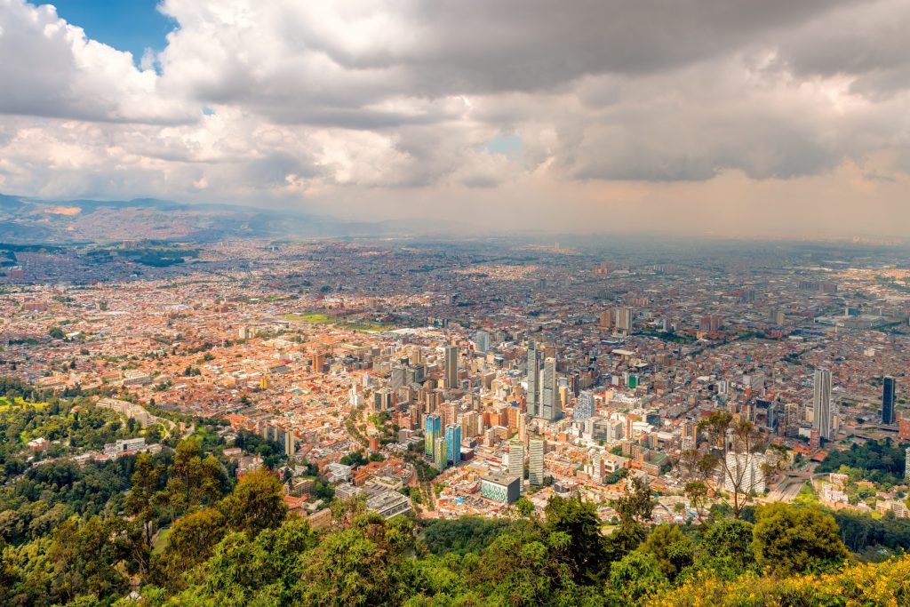 Aerial view of Bogota, Colombia, featuring a dense cityscape under cloudy skies, framed by green trees in the foreground.