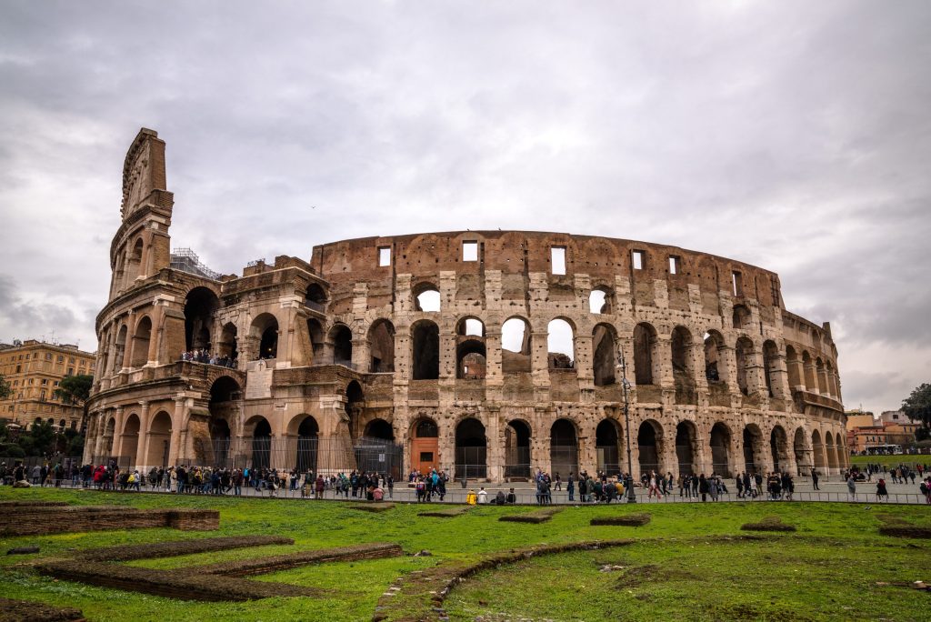 The Colosseum in Rome, Italy, stands under a cloudy sky. Tourists walk around the ancient amphitheater, surrounded by green grass.