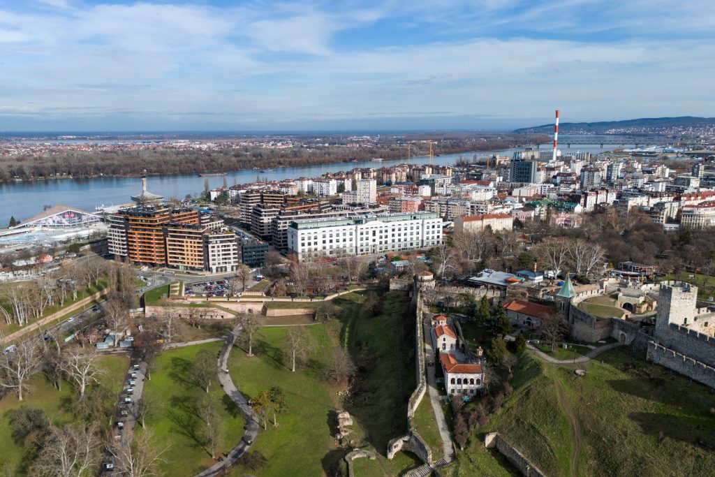 Aerial view of Belgrade, Serbia featuring the fortress, river, modern buildings, and green parkland under a blue sky.