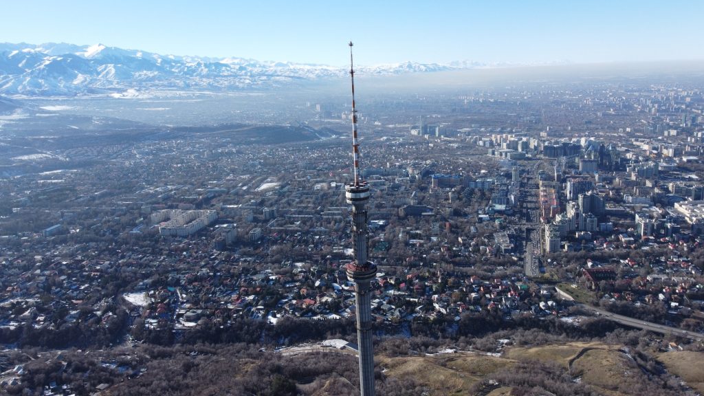 Tall Almaty TV Tower rises above city, with snow-covered mountains in background under a clear blue sky.