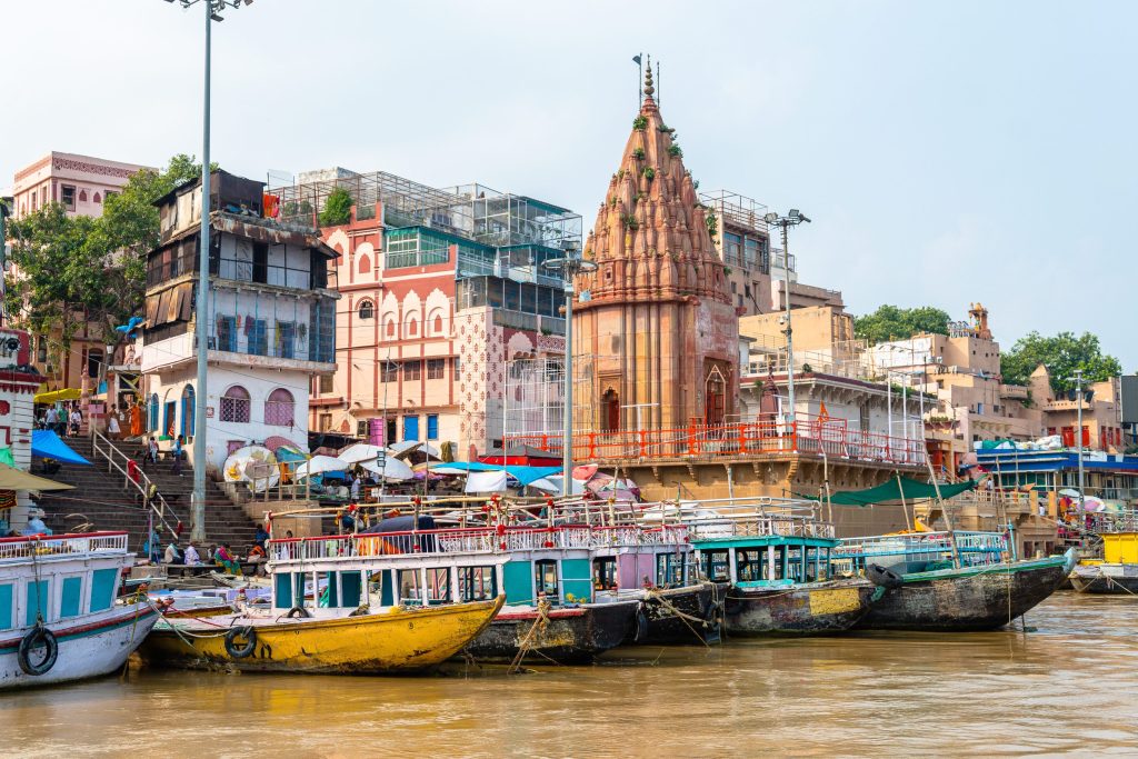 Colorful boats line the Ganges River in Varanasi, India, with a temple and buildings in the background.