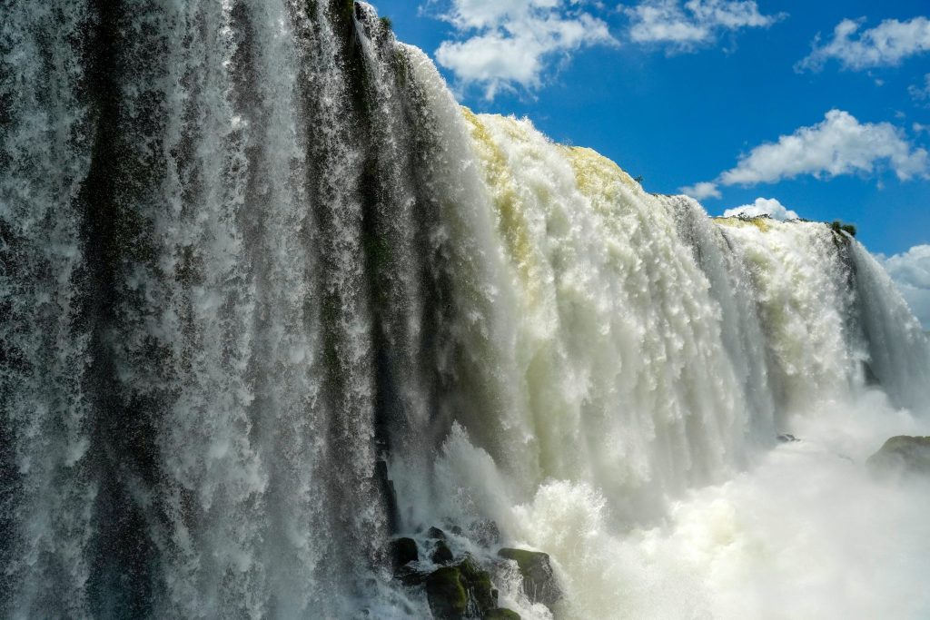Powerful waterfalls cascade over a cliff into a misty pool, blue sky above.