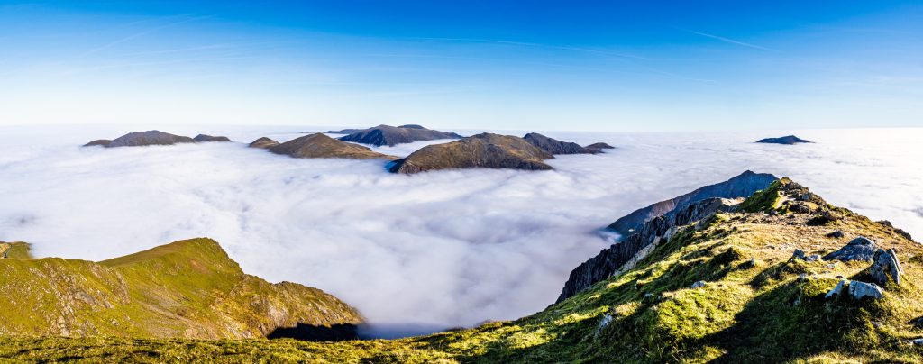 Panoramic view of mountain peaks rising above a sea of clouds in Snowdonia, Wales, under a clear blue sky.