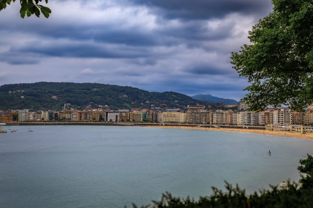 Coastal city view of San Sebastian, Spain with buildings, beach, ocean, and cloudy sky. Green trees frame the scene.