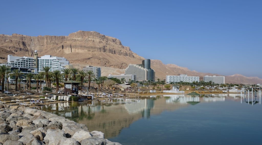 A scenic view of Dead Sea hotels and resorts with mountains in the background. The water is calm and blue.