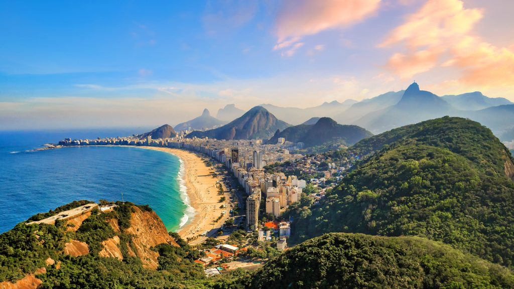 Aerial view of Rio de Janeiro, Brazil, featuring Copacabana Beach, mountains, and cityscape under a blue sky.