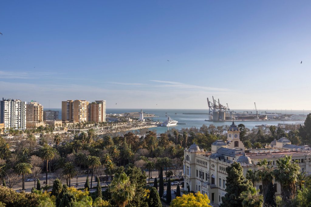 Aerial view of Malaga, Spain, featuring modern buildings, port with cranes, and historic palace under a blue sky.
