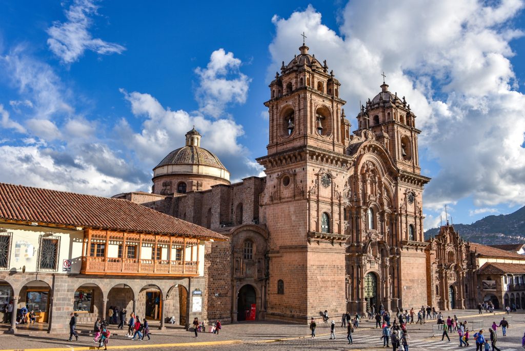Cusco Cathedral in Peru, with people walking in the Plaza de Armas under a cloudy blue sky.