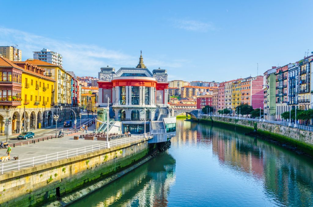 Bilbao, Spain cityscape with colorful buildings lining a river, clear blue sky. People walk along the riverfront.