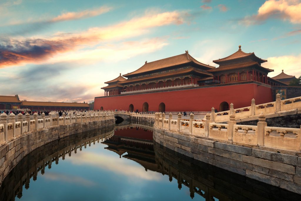 The Forbidden City in Beijing, China, featuring red walls, golden roofs, and a reflective moat under a colorful sunset sky.