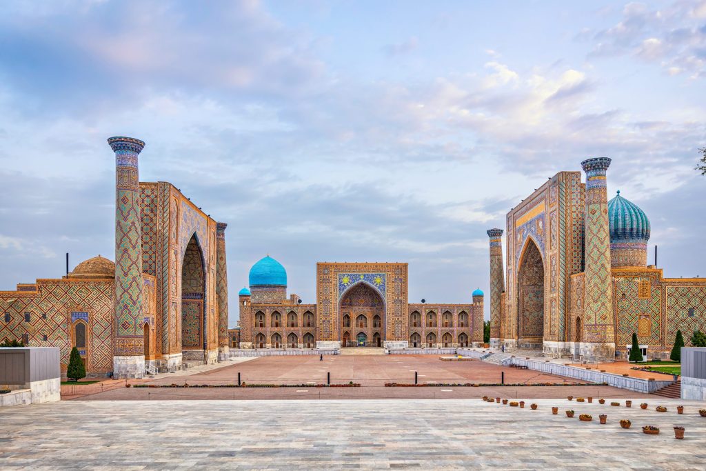 Wide shot of Registan Square in Samarkand, Uzbekistan, featuring ornate tilework, blue domes, and minarets under a cloudy sky.