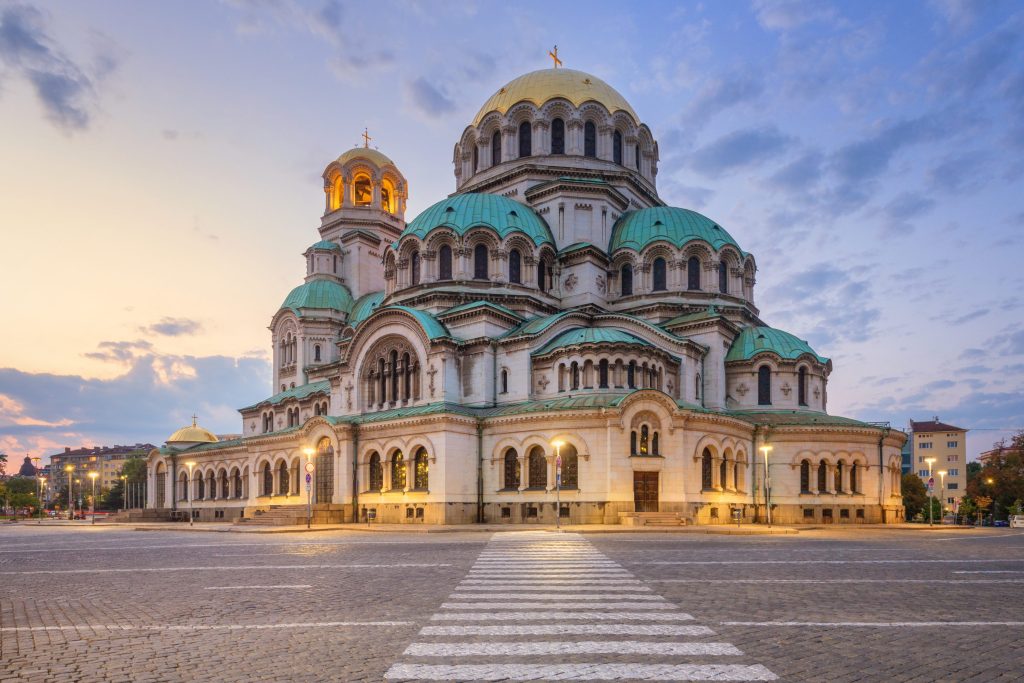 Grand cathedral with gold and green domes stands in a cobblestone square under a cloudy sky.