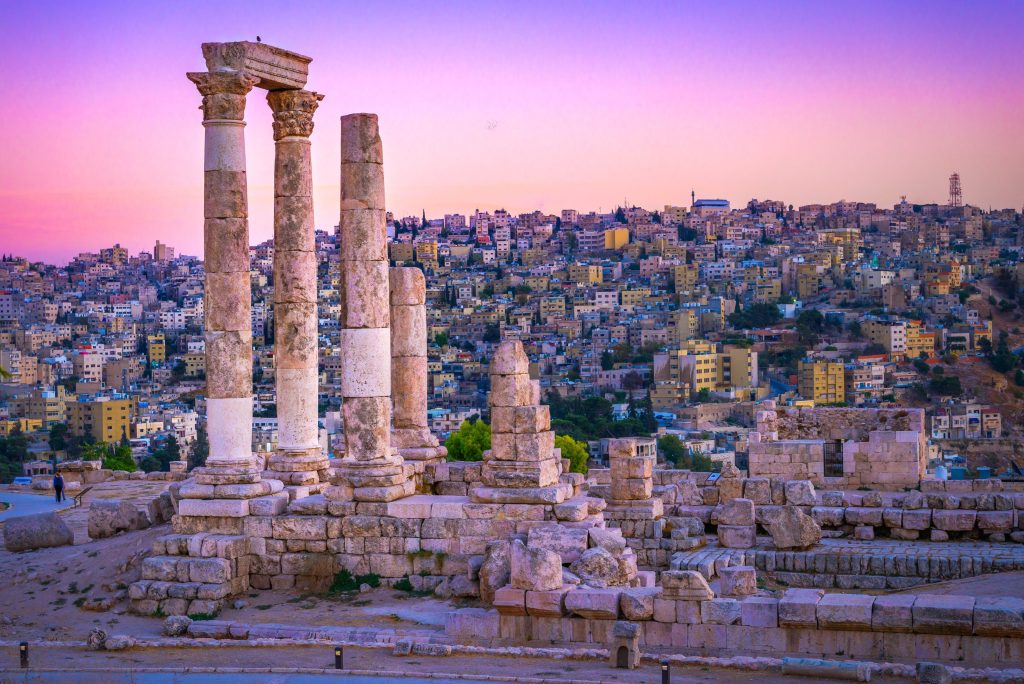 Ancient Roman columns stand in Amman, Jordan, with a cityscape backdrop under a pink and purple sunset.