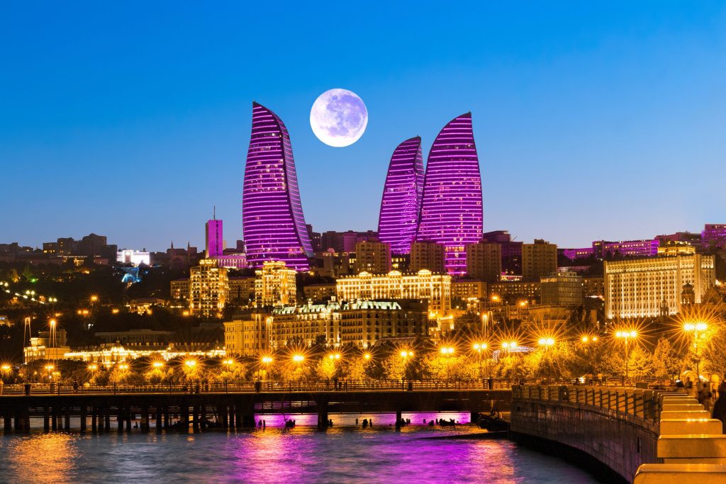 Night view of Baku, Azerbaijan skyline with illuminated Flame Towers, buildings, and waterfront reflecting purple light.