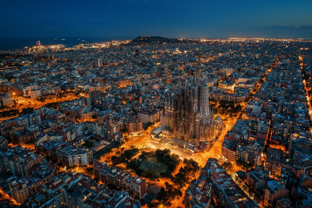 Aerial view of Barcelona at night. The Sagrada Familia cathedral is illuminated, surrounded by city lights and dark blue sky.