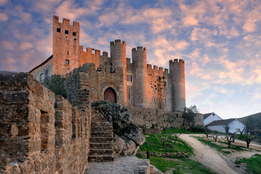 Stone castle with towers and crenellations, bathed in warm sunset light, overlooks a path and white buildings in Óbidos, Portugal.