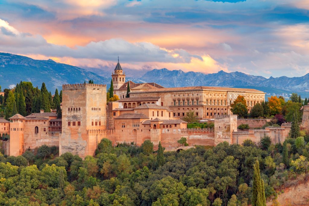 Granada's Alhambra Palace at sunset, a sprawling complex of reddish buildings, towers, and lush green trees against a backdrop of mountains.