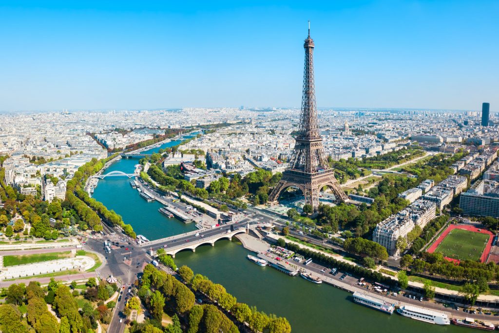 Aerial view of Paris featuring the Eiffel Tower, Seine River, bridges, buildings, and green spaces under a clear blue sky.