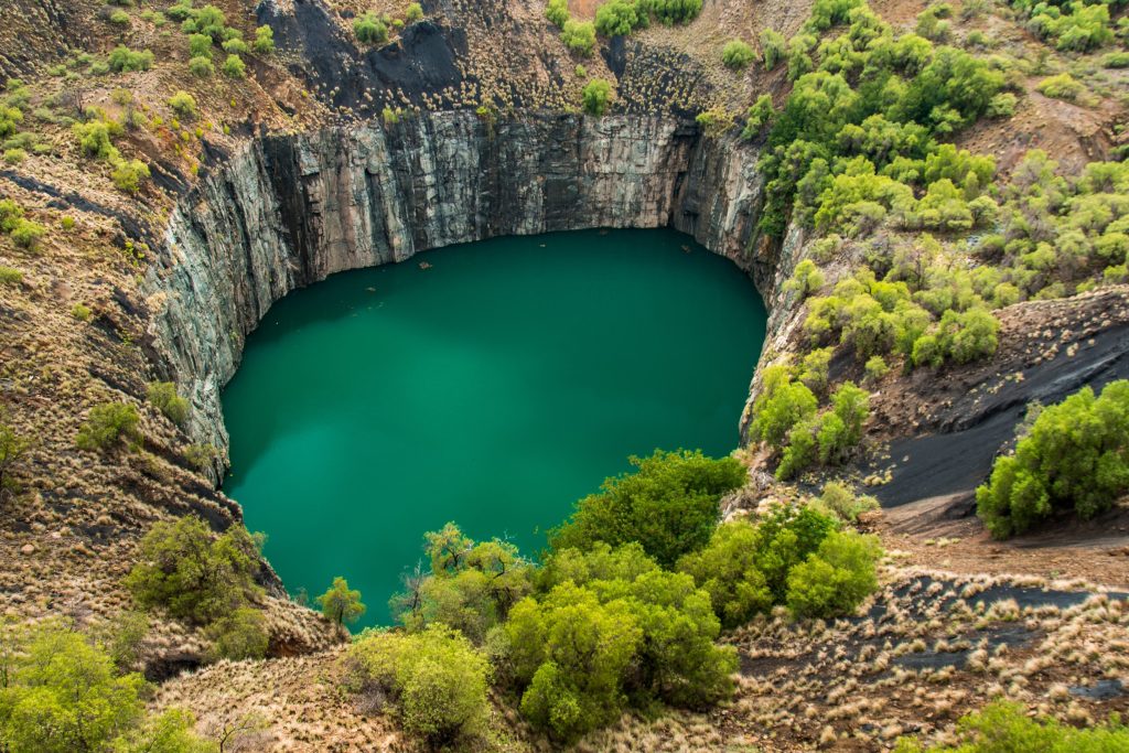 Aerial view of a large turquoise lake within a deep, rocky open-pit mine surrounded by sparse vegetation.