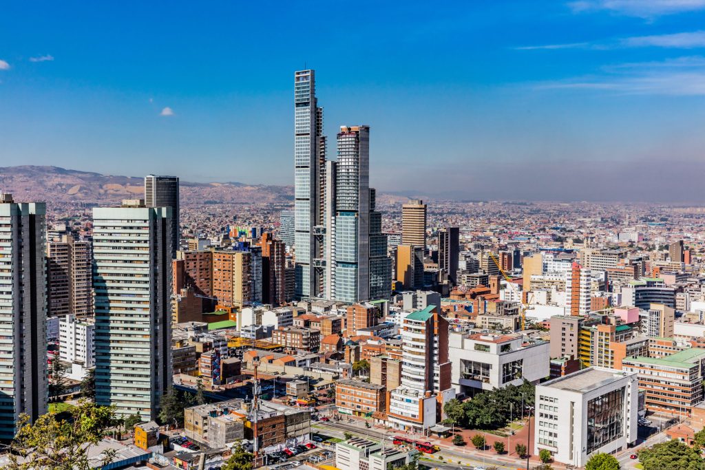 Wide cityscape view of Bogota, Colombia, featuring modern high-rise buildings under a bright blue sky.