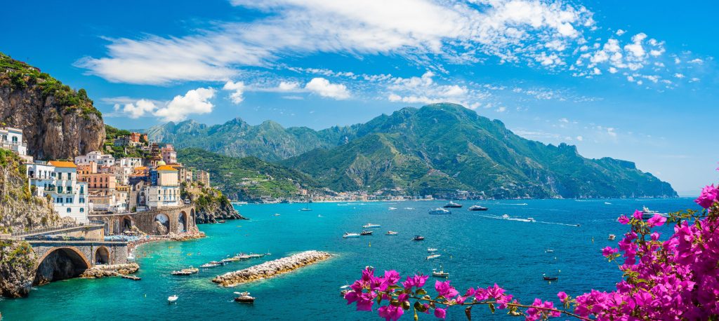 Colorful Amalfi Coast town, Italy, with turquoise sea, mountains, and vibrant pink bougainvillea flowers in foreground.