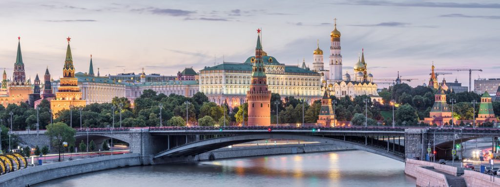 Moscow Kremlin and cityscape at dusk, featuring golden domes, towers, bridge, and river reflections.