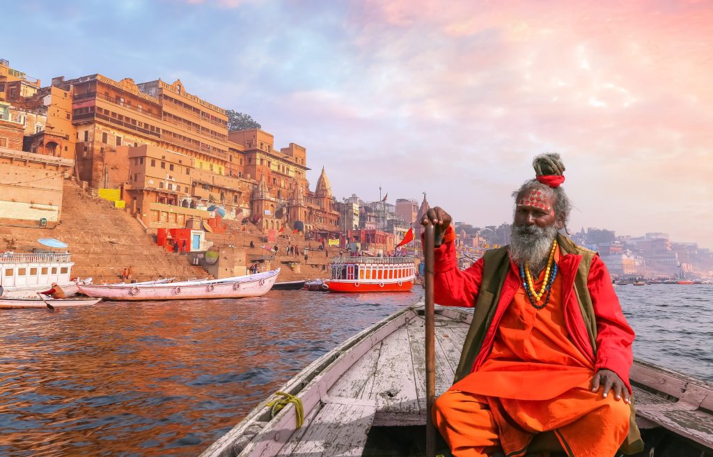 A sadhu sits in a boat on the Ganges River, Varanasi, India, with colorful buildings in the background.