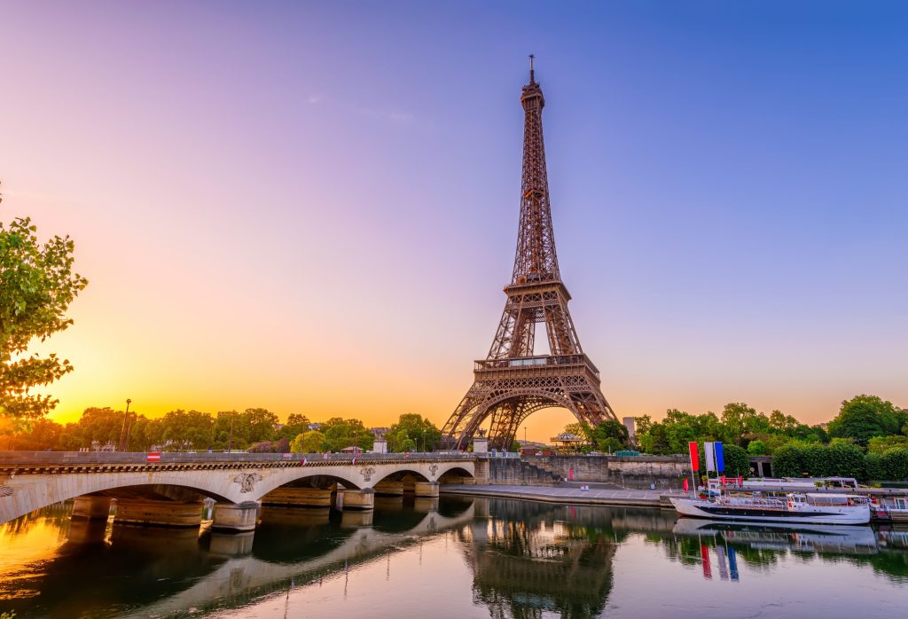 Paris cityscape at sunset featuring the Eiffel Tower, bridge over the Seine, and tour boat.