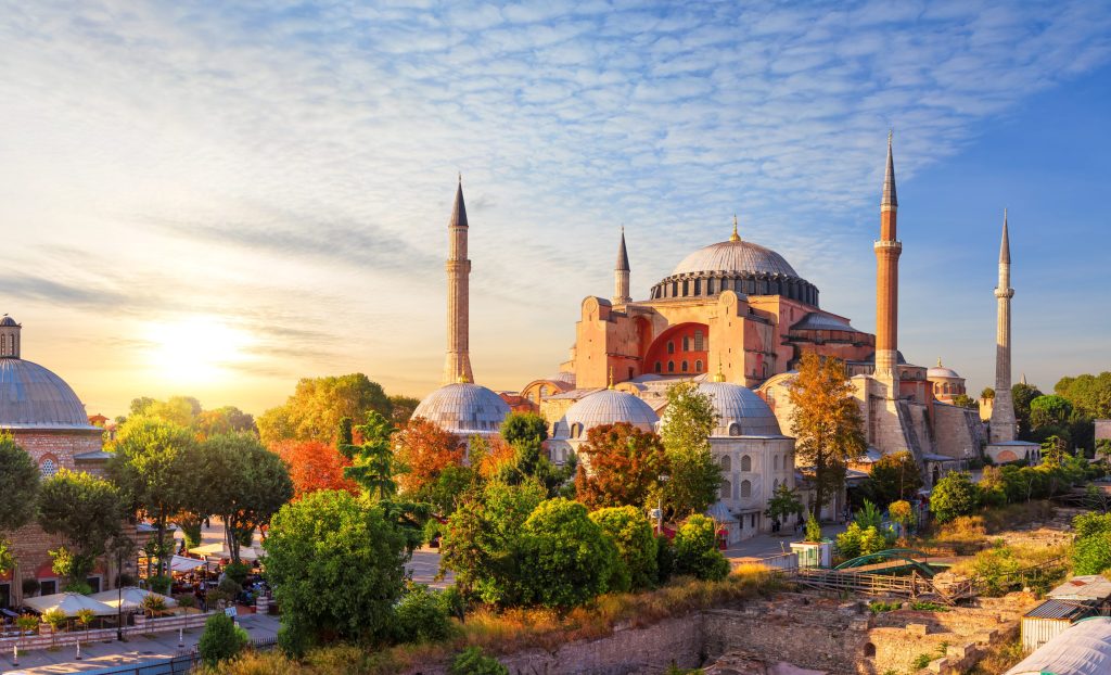Hagia Sophia in Istanbul, Turkey, with minarets and domes, bathed in golden sunrise light, surrounded by trees.