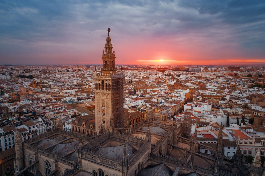 Aerial view of Seville Cathedral tower rising above the city at sunset, orange glow on horizon, cloudy sky.