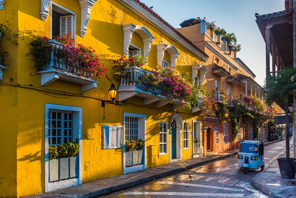 Bright yellow buildings line a wet Cartagena street, adorned with flower-filled balconies, and a blue tuk-tuk drives by.