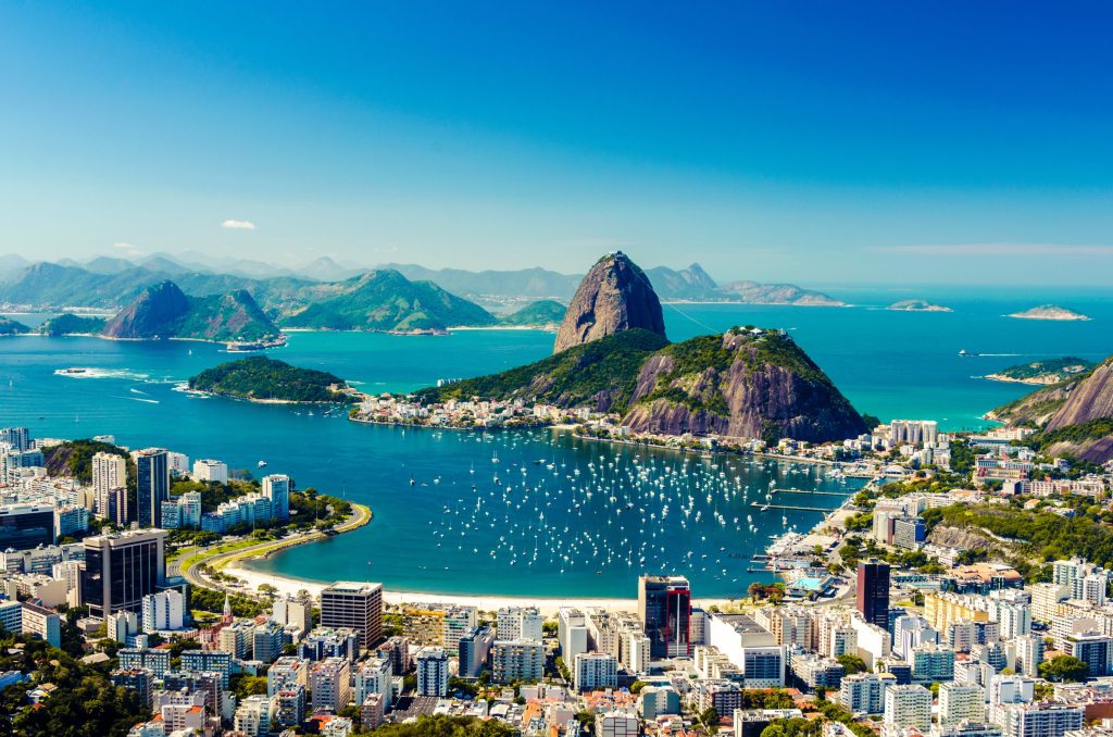 Aerial view of Rio de Janeiro, Brazil featuring Sugarloaf Mountain, Guanabara Bay, and city buildings under a clear blue sky.