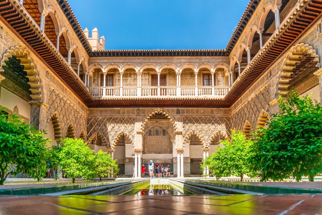 Courtyard of Alcázar of Seville features intricate Moorish architecture, lush greenery, and a tranquil reflecting pool. People are walking around.