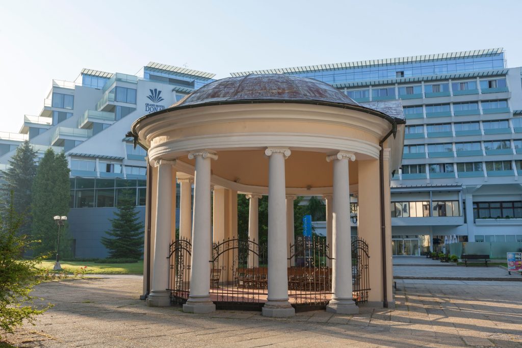 A circular pavilion with columns and a dome roof stands in front of a large hotel.