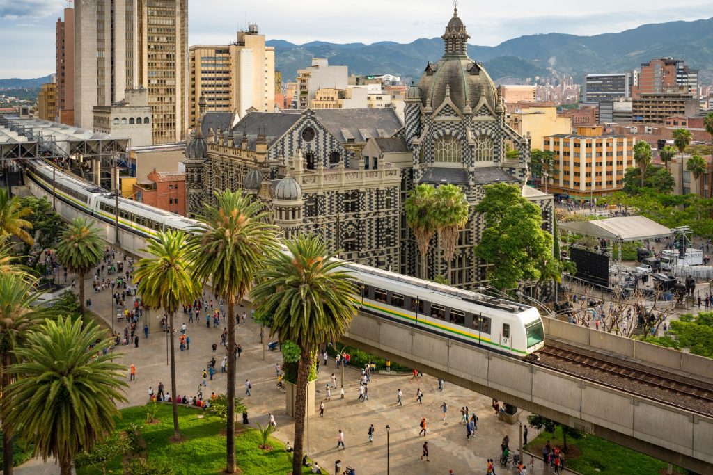 Elevated train passes Rafael Uribe Uribe Palace of Culture in Medellín, Colombia, with people walking below.