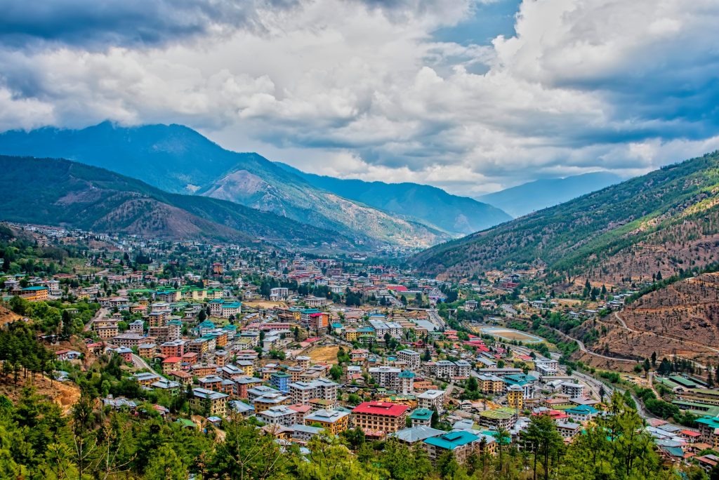 High angle view of Thimphu, Bhutan, nestled in a green valley surrounded by blue mountains under cloudy skies.